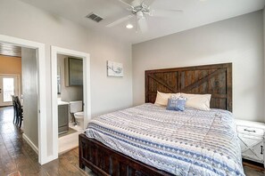 Une chambre à coucher avec un grand lit, une tête de lit en bois, un ventilateur de plafond et une salle de bain visible à travers une porte ouverte.