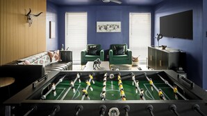 A room with a foosball table, two green armchairs, a sofa, a ceiling fan, and a television on the wall. - Sport Court Private Heated Pool Mini Golf More (Glendale)