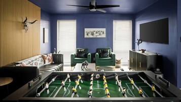 A room with a foosball table, two green armchairs, a sofa, a ceiling fan, and a television on the wall.