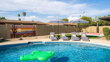 A swimming pool with lounge chairs and umbrellas, a mural on the wall, and a basketball hoop.