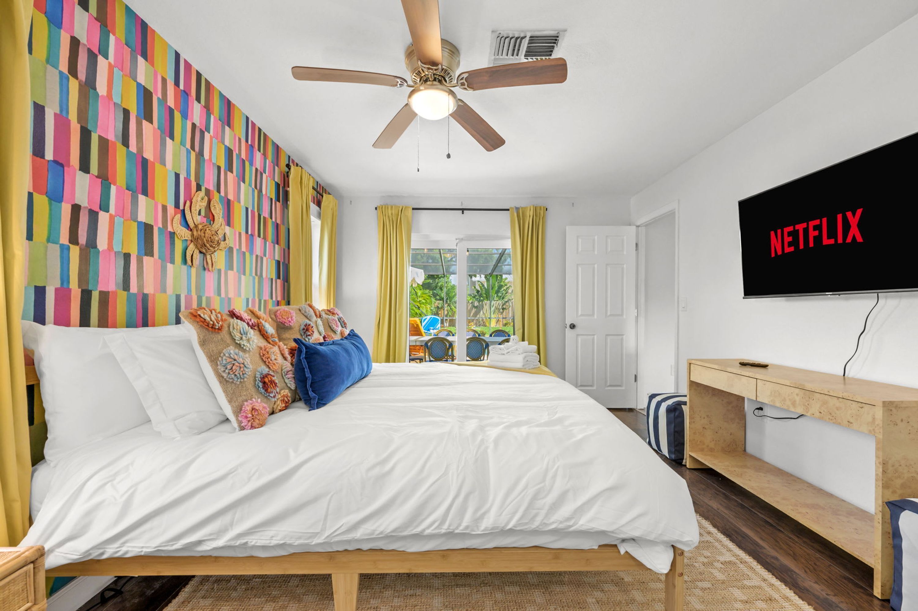 A hotel room with a bed, a ceiling fan, a TV mounted on the wall, and a view of the outdoors through a window.