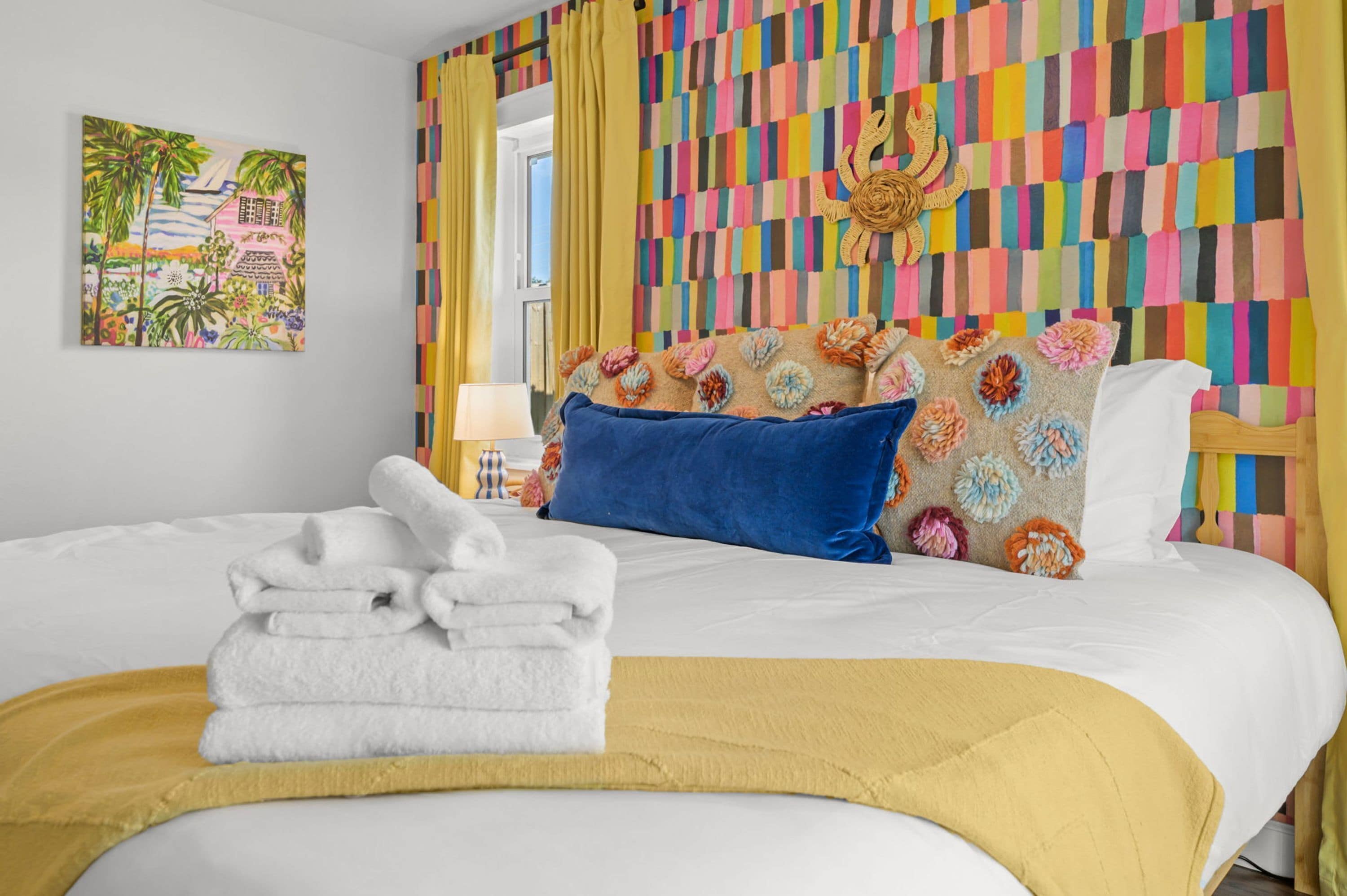 Una habitación de hotel con una cama, almohadas, una pila de toallas, una pared de colores y un cuadro.