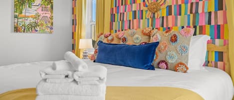 Una habitación de hotel con una cama, almohadas, una pila de toallas, una pared de colores y un cuadro.
