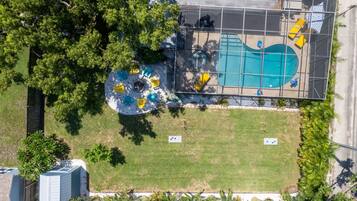 An aerial view of a swimming pool area with a fenced deck, surrounded by trees and grass.
