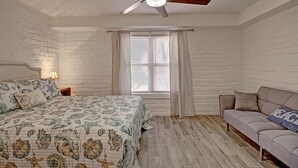 A bedroom with a bed, a ceiling fan, a window with curtains, a nightstand, and a sofa. - A Salty Day Getaway Beach Boardwalk Large Pool (Port Aransas)