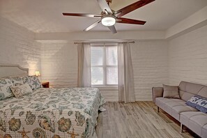A bedroom with a bed, a ceiling fan, a window with curtains, a nightstand, and a sofa. - A Salty Day Getaway Beach Boardwalk Large Pool (Port Aransas)