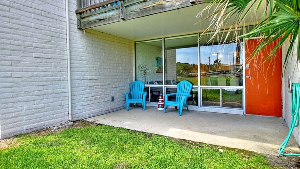 A patio with two chairs and a table, a glass door leading to a covered area, and a palm tree. - A Salty Day Getaway Beach Boardwalk Large Pool (Port Aransas)