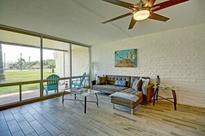 A spacious living area with a large window, a ceiling fan, and a painting on the wall. - A Salty Day Getaway Beach Boardwalk Large Pool (Port Aransas)