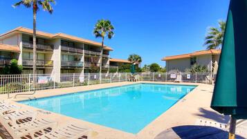 A swimming pool with lounge chairs, a fenced area, and a building with balconies.