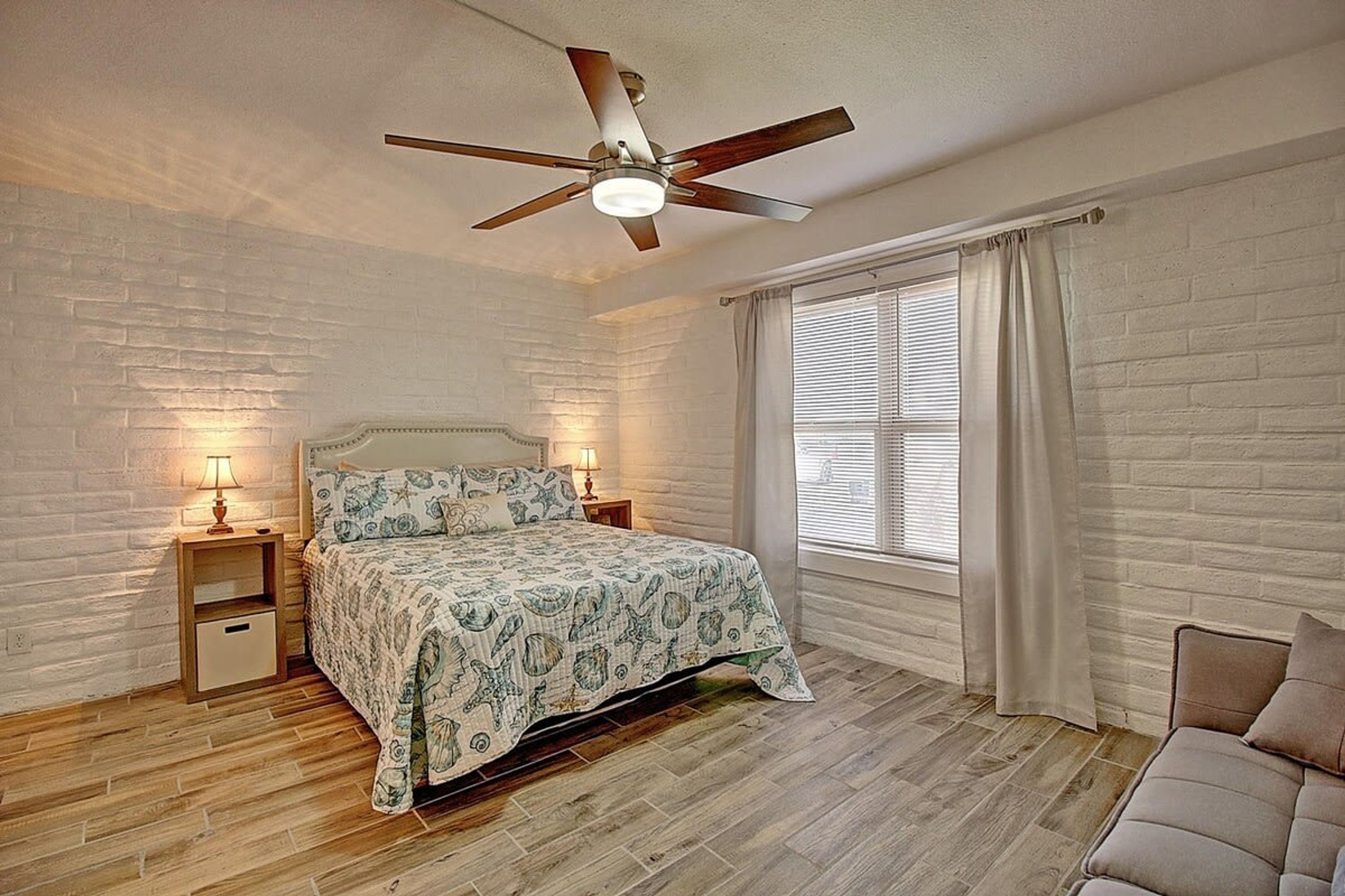 Une chambre à coucher avec un lit, des tables de chevet, un ventilateur de plafond, une fenêtre avec des rideaux et un mur en briques.