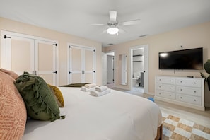 Ein Schlafzimmer mit einem Bett, einem Deckenventilator, einem Fernseher und einem durch eine offene Tür einsehbaren Badezimmer.