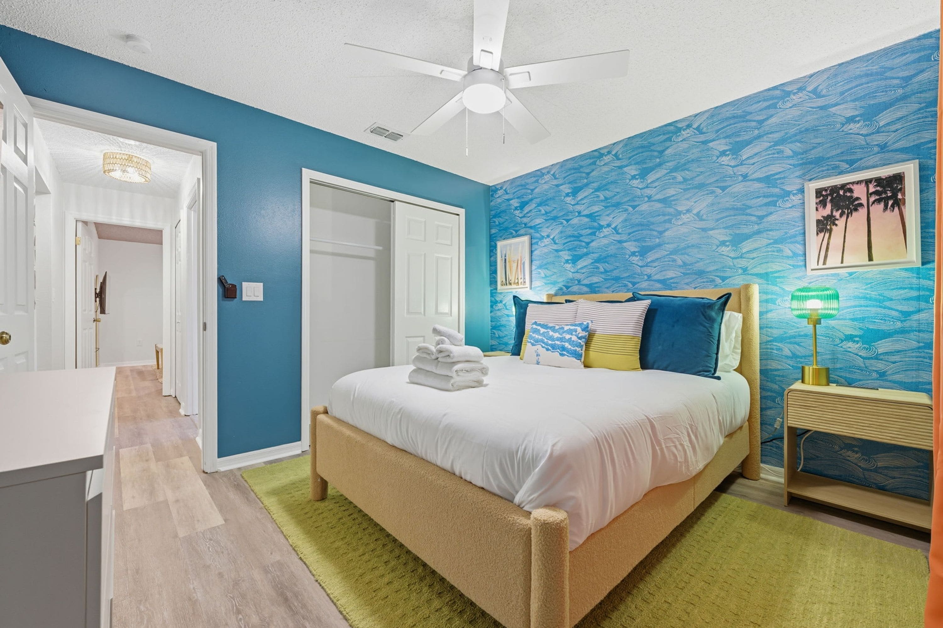 Une chambre à coucher comprenant un lit, une table de chevet, un ventilateur de plafond et un mur orné d’un motif bleu.