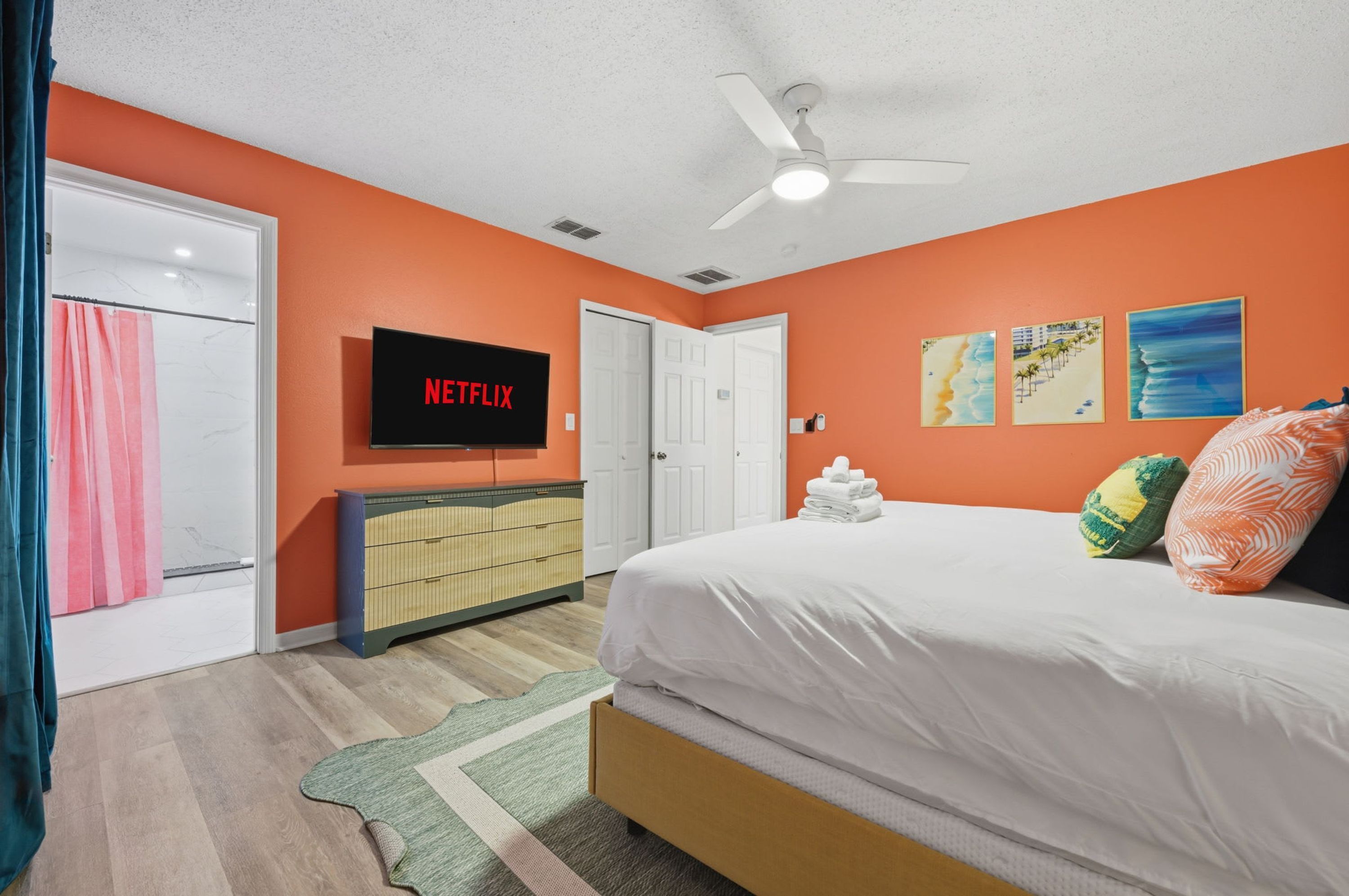 A bedroom with a bed, a TV mounted on the wall, a ceiling fan, and artwork on the wall.