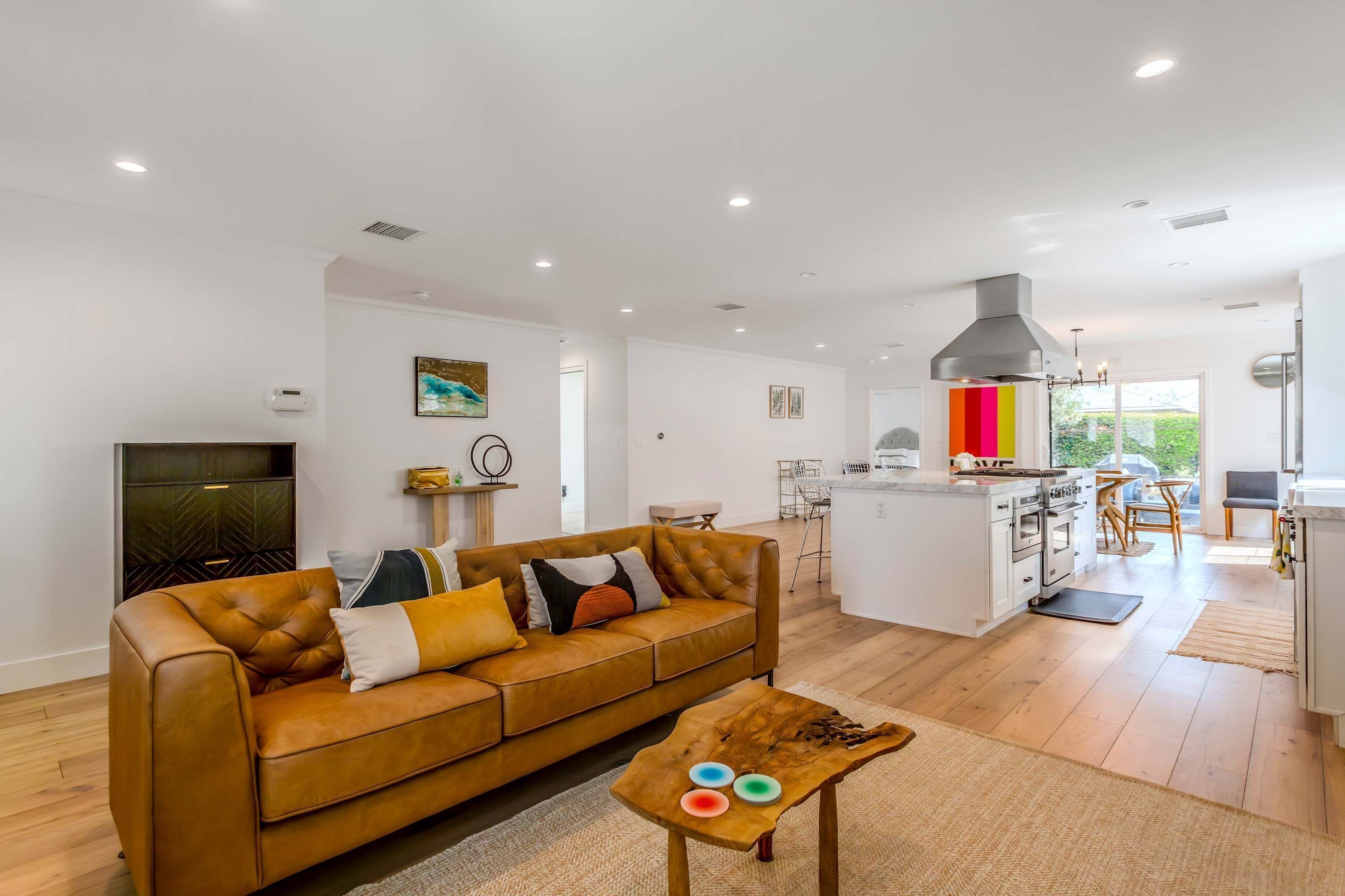 Uma sala de estar moderna com um sofá de couro, uma mesa de centro com pratos coloridos e uma cozinha com ilha central e eletrodomésticos de aço inoxidável.