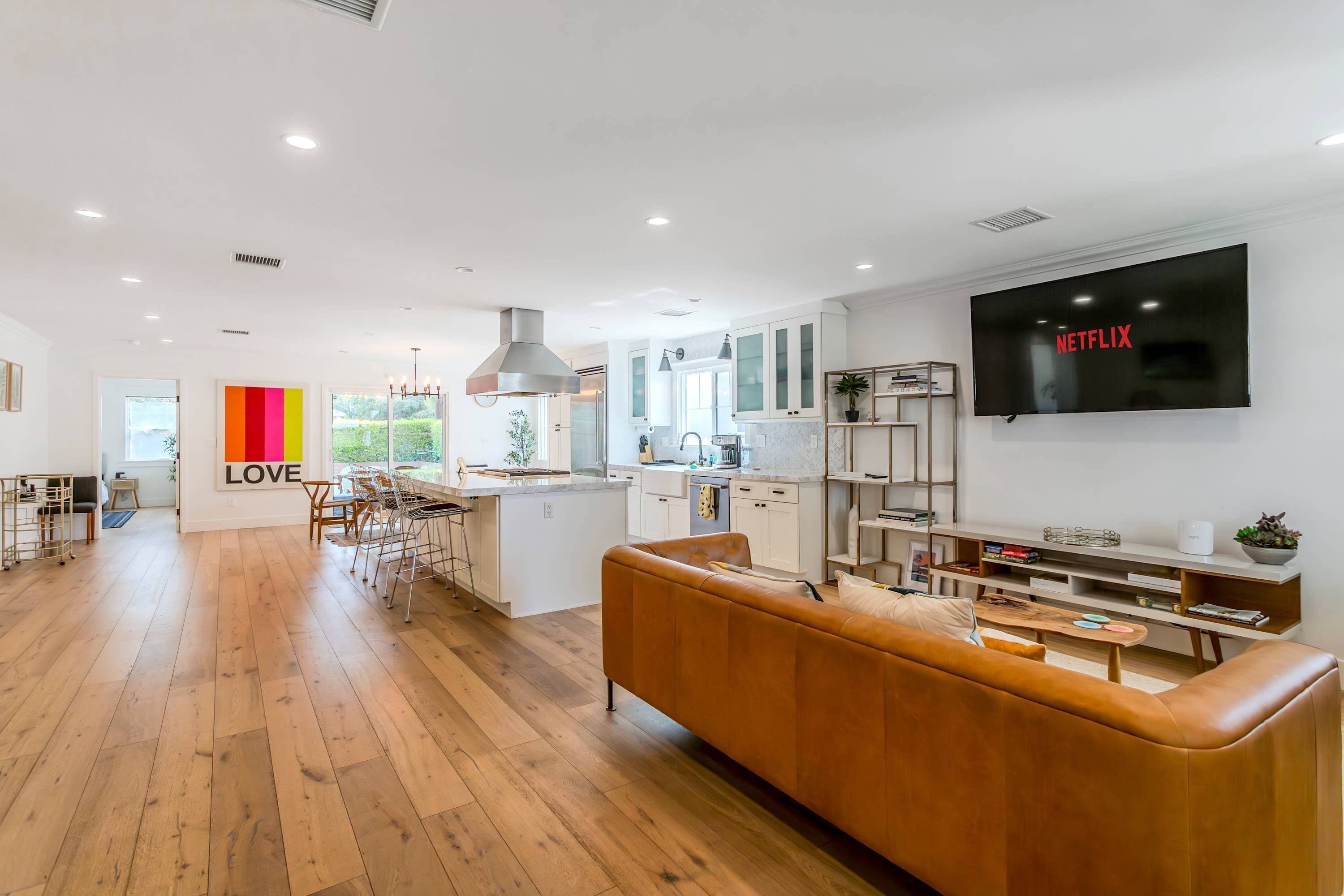 Uma cozinha moderna com uma ilha central, uma área de jantar com um banquinho de bar e uma sala de estar com uma televisão grande.