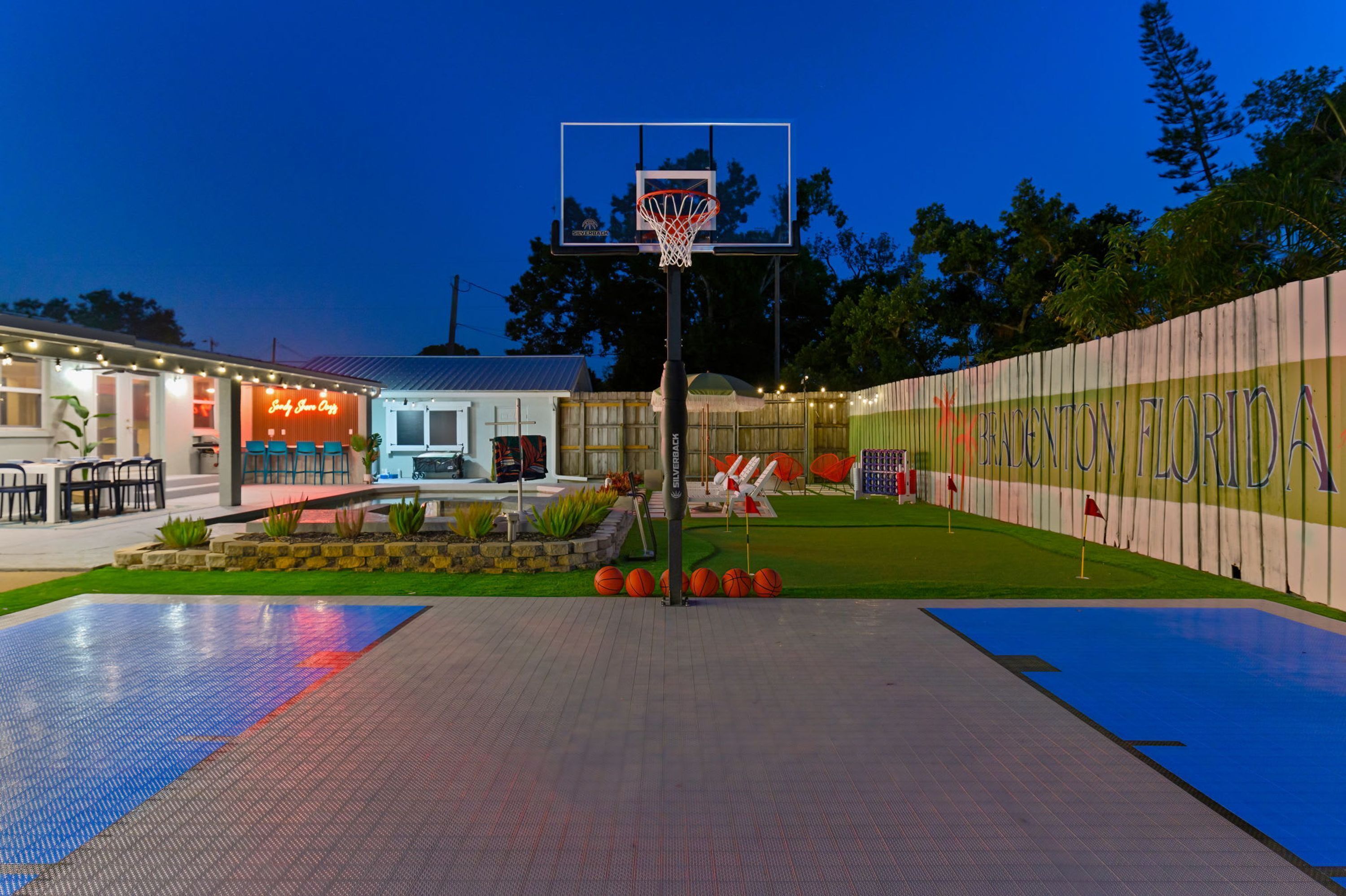 A basketball court with a hoop, surrounded by a fence with a sign, and a building with outdoor seating.