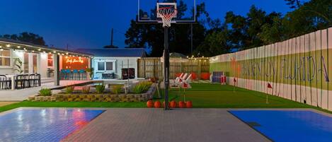 A basketball court with a hoop, surrounded by a fence with a sign, and a building with outdoor seating.