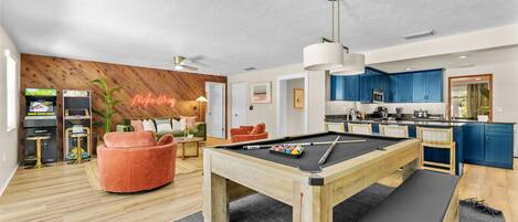 A modern living space with a pool table, a neon sign, a kitchen, and a wooden wall.