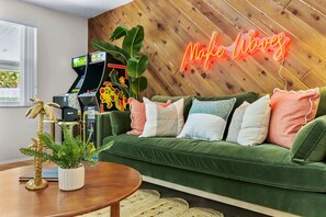 Un canapé vert agrémenté de plusieurs coussins, un panneau lumineux néon, une console de jeux et une plante en pot.