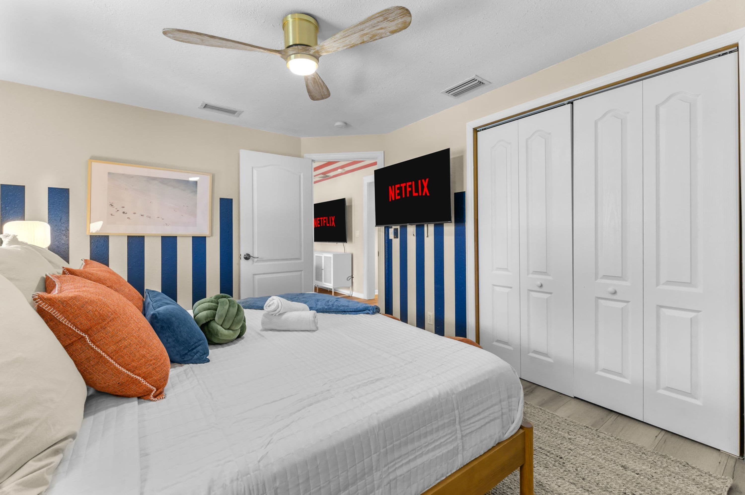 A bedroom with a bed, pillows, a ceiling fan, a framed picture, a television, and a wardrobe.