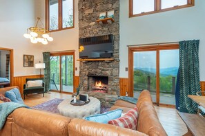 A spacious living room with a stone fireplace, leather sofas, a television mounted on the wall, and large windows offering a view of the outdoors. - Sleeps 20 Hot Tub Outdoor Theater Fire Pit Games (Fairview)