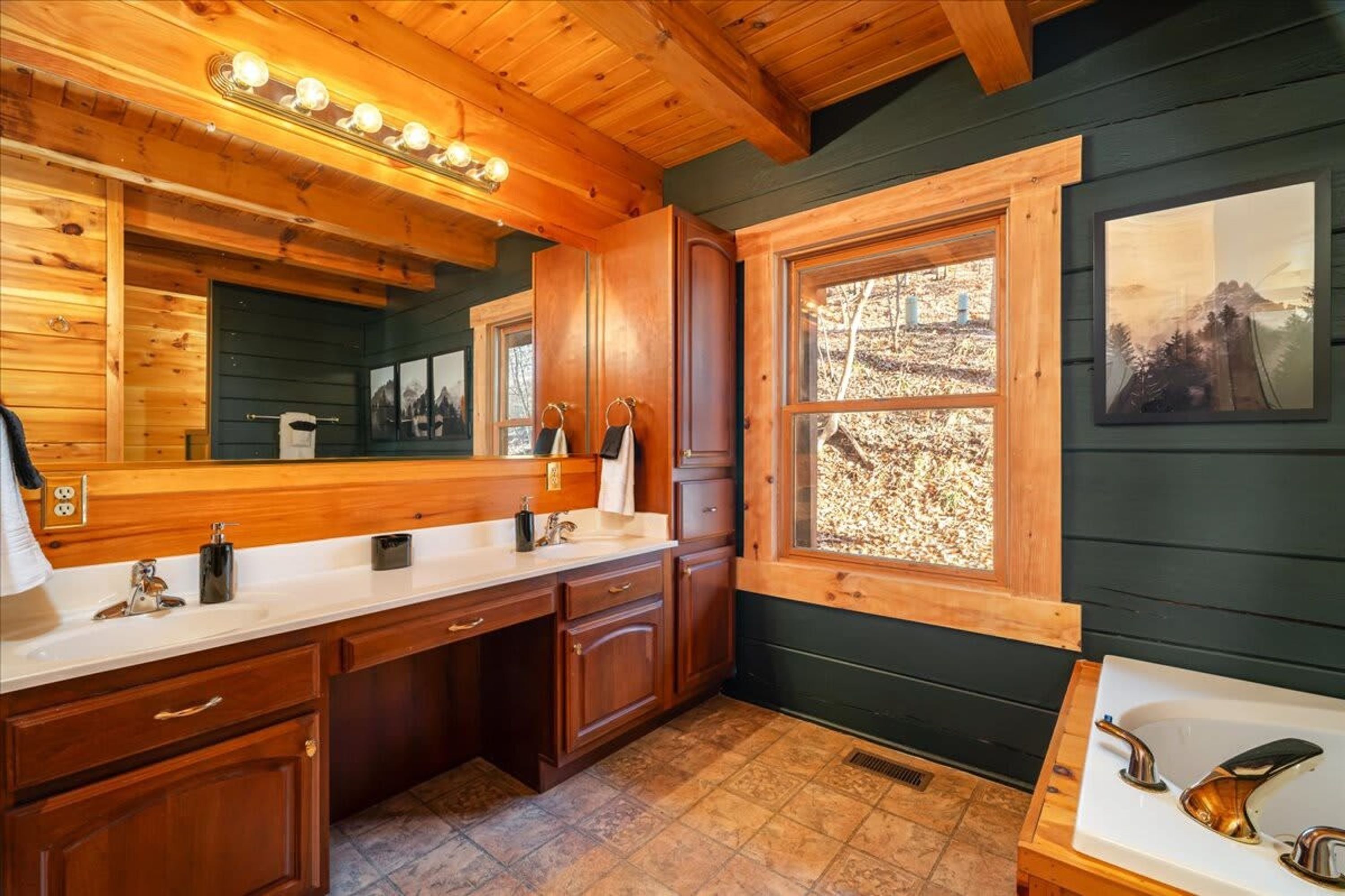 Ein Badezimmer in einer Holzhütte mit einem großen Spiegel, Holzausbau, einem Waschbecken und einem Fenster mit Blick auf Bäume.