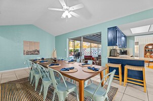 Comedor con una mesa dispuesta para cuatro personas, sillas azul claro, ventilador de techo y vista a un área exterior a través de un vidrio.