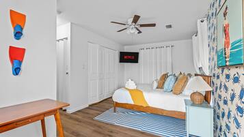 Un dormitorio con cama, televisión, ventilador de techo y baño visible a través de una puerta abierta.