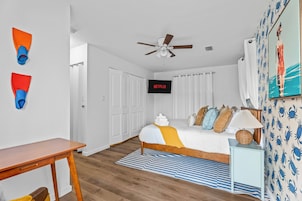 Un dormitorio con cama, televisión, ventilador de techo y baño visible a través de una puerta abierta.