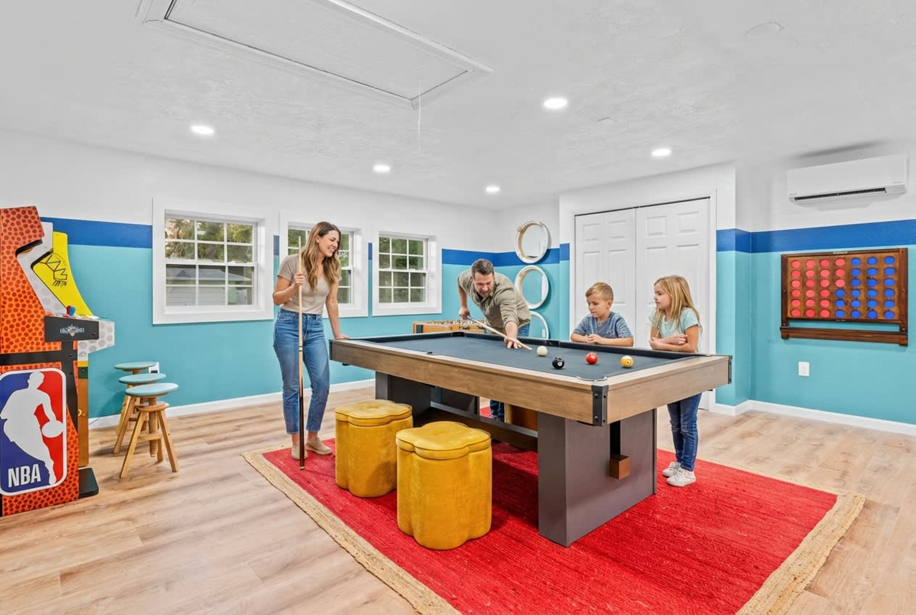 A pool table in a room with a red carpet, wooden stools, and a basketball arcade game.