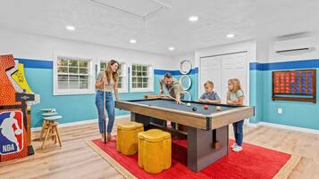 A pool table in a room with a red carpet, wooden stools, and a basketball arcade game.