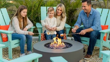 Una familia de cuatro sentada en sillas Adirondack turquesas alrededor de un pozo de fuego circular con fuego encendido.