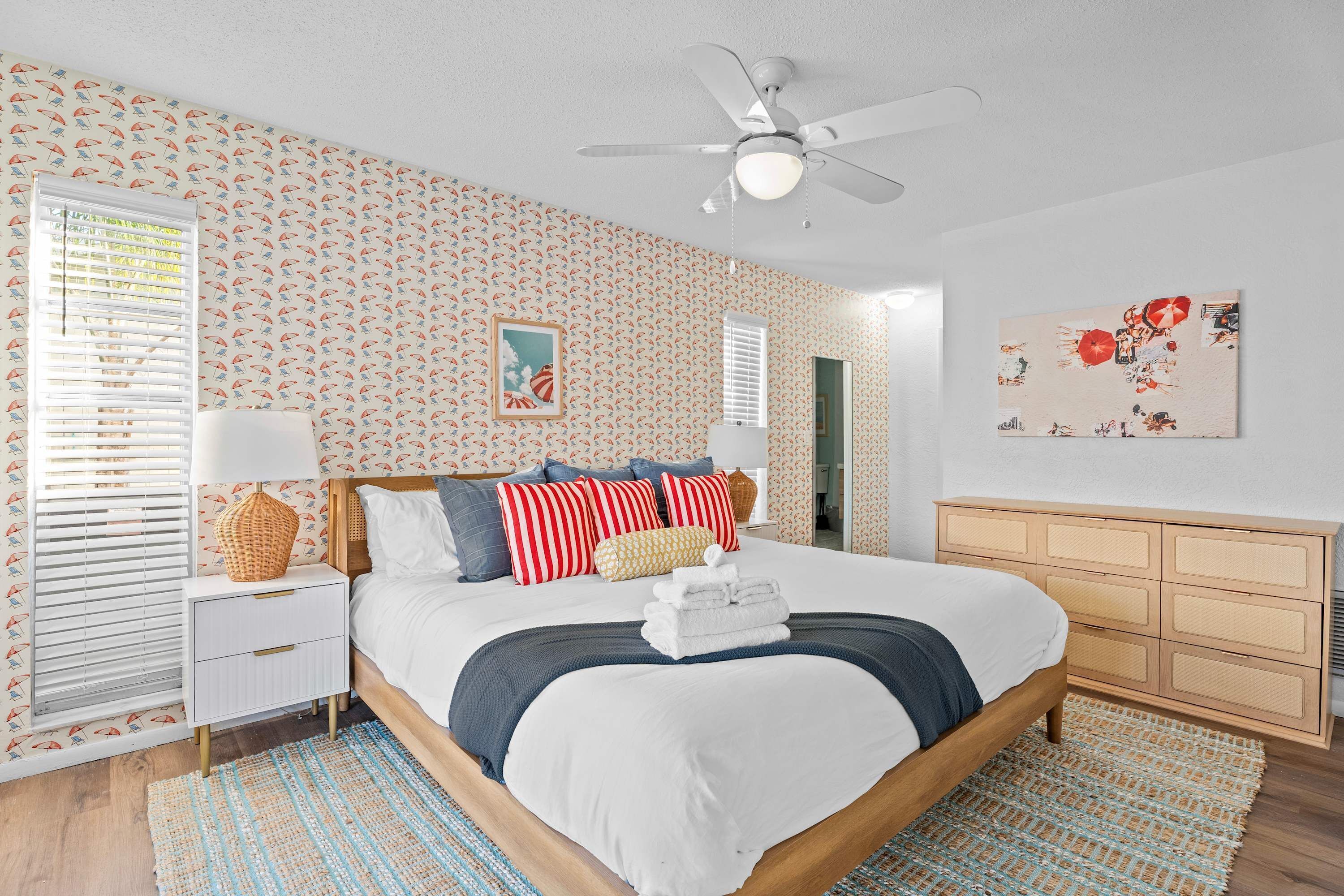A bedroom with a bed, bedside tables, a dresser, a ceiling fan, and patterned wallpaper.