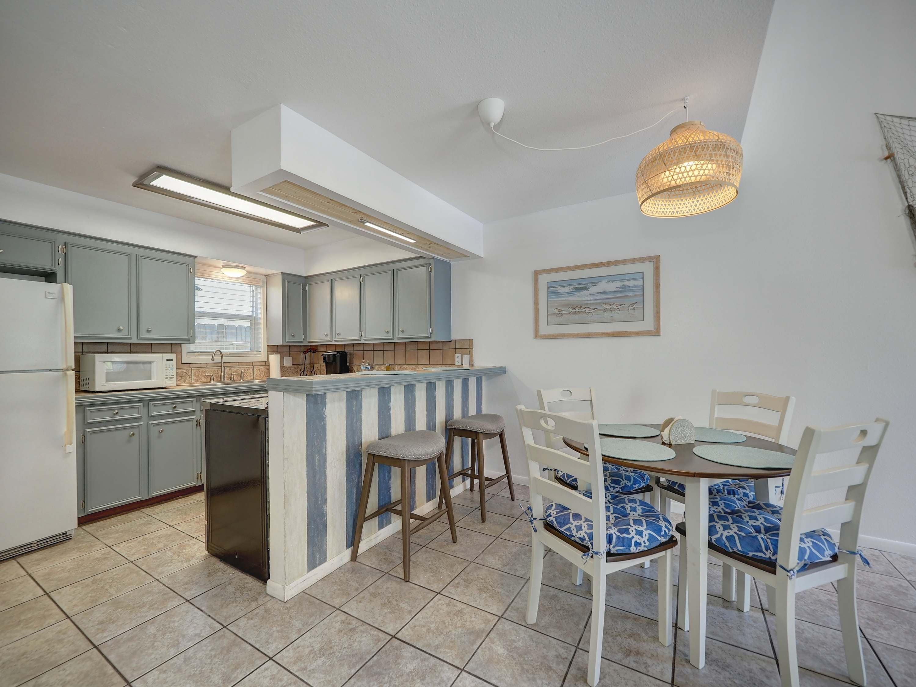Een keuken met een inbouwkookeiland, grijze kastjes en een eethoek met witte stoelen en blauwe kussens.