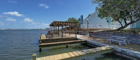 Un muelle de madera que se adentra en un cuerpo de agua, con una zona de estar cubierta y un edificio al fondo.