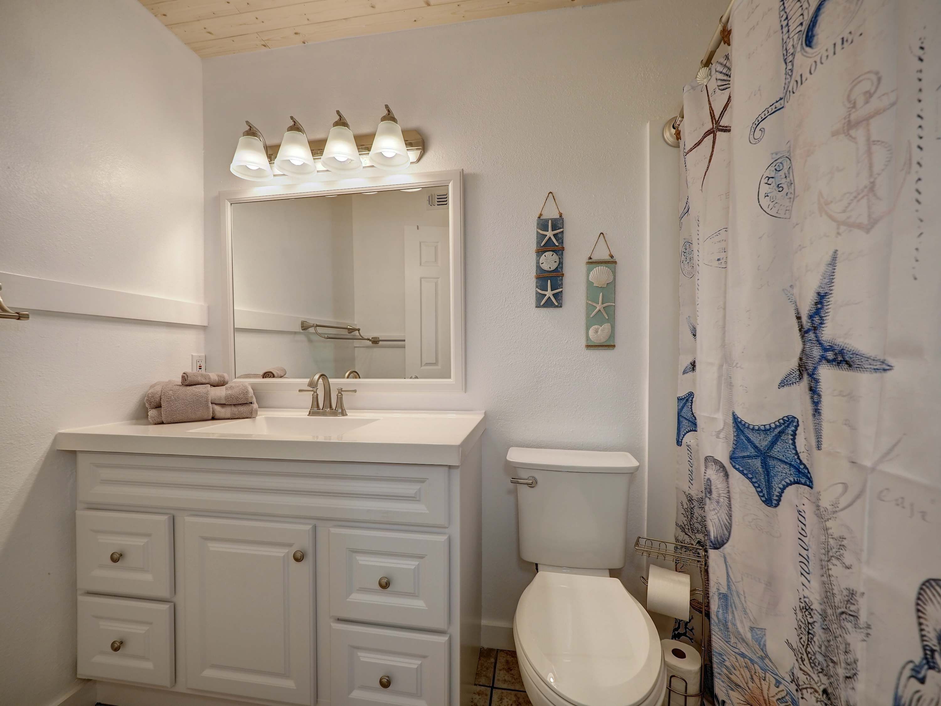 Ein Badezimmer mit weißem Waschtisch, einem Waschbecken, einem Spiegel und einer Toilette. Auf dem Waschtisch liegen Handtücher, der Duschvorhang ist mit maritimen Motiven versehen und eine Wandleuchte ist angebracht.