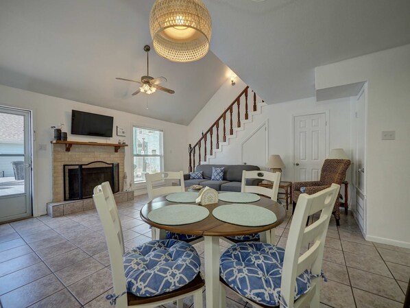 A dining area with a round table, chairs with patterned cushions, a ceiling fan, a fireplace, and a staircase leading upstairs.