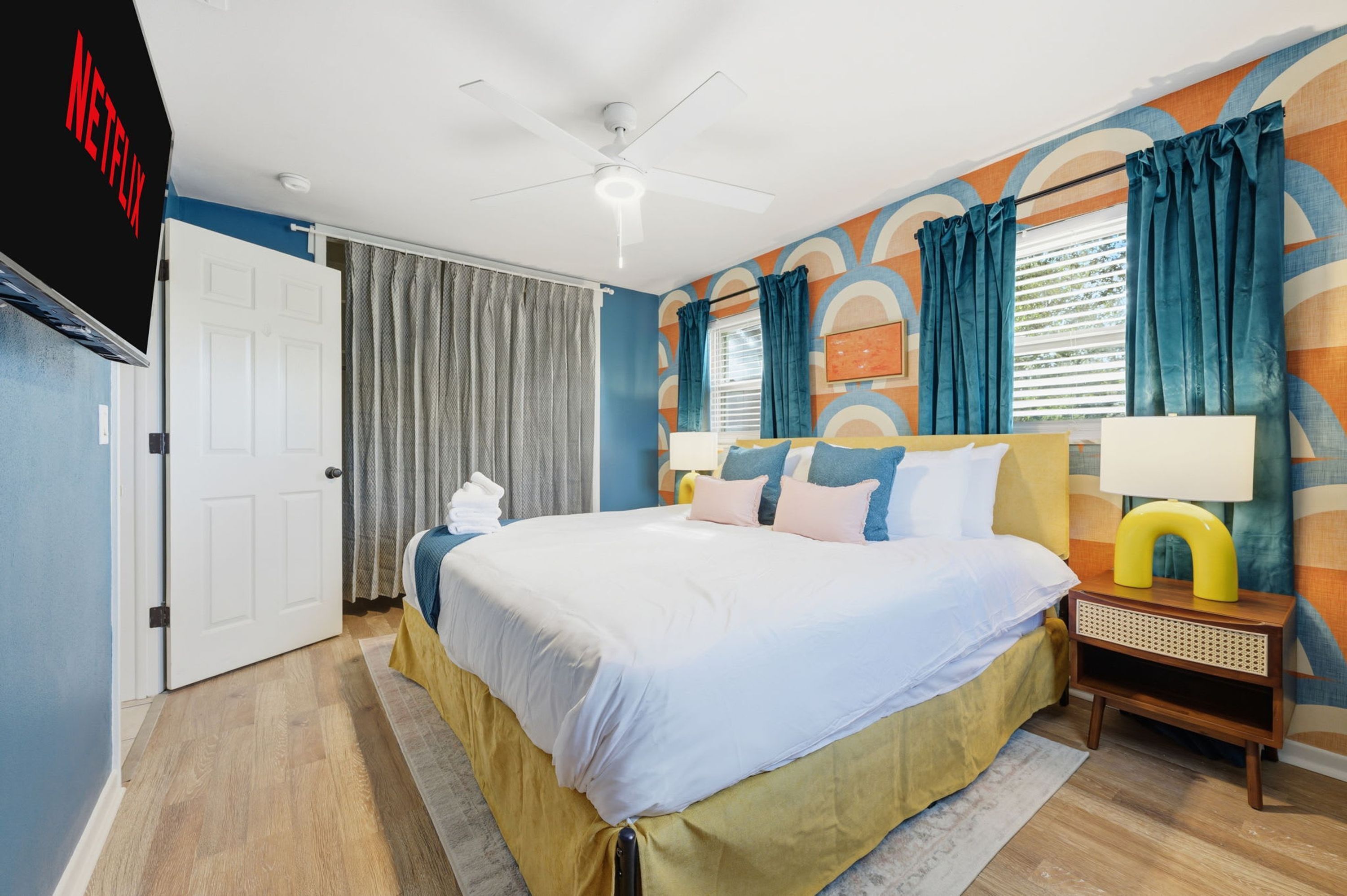 Quarto de hotel com cama, mesa de cabeceira com abajur, ventoinha no teto e parede com padrão geométrico.