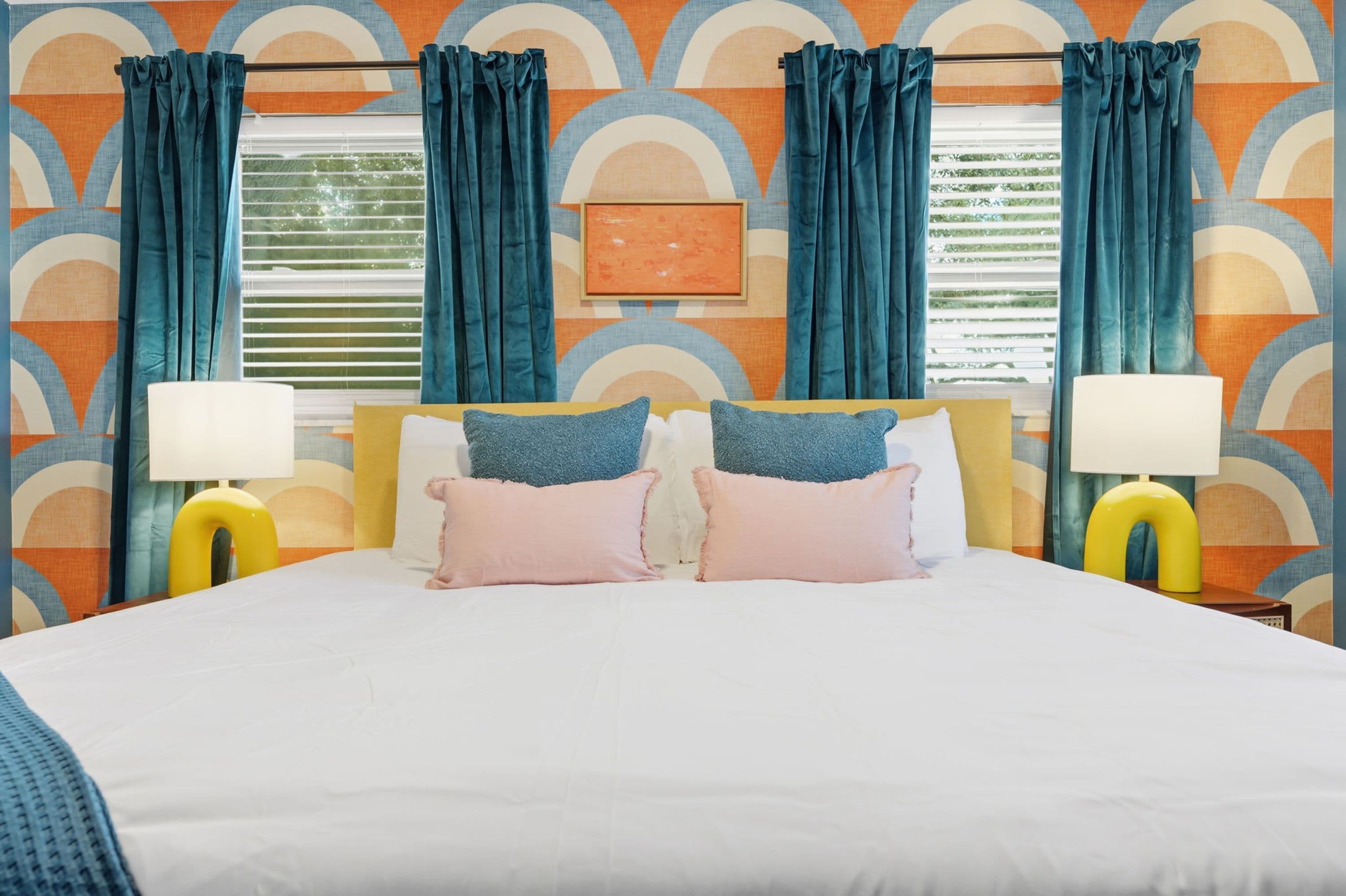 Una habitación de hotel con una cama, dos lámparas de noche y un ventanal con cortinas.