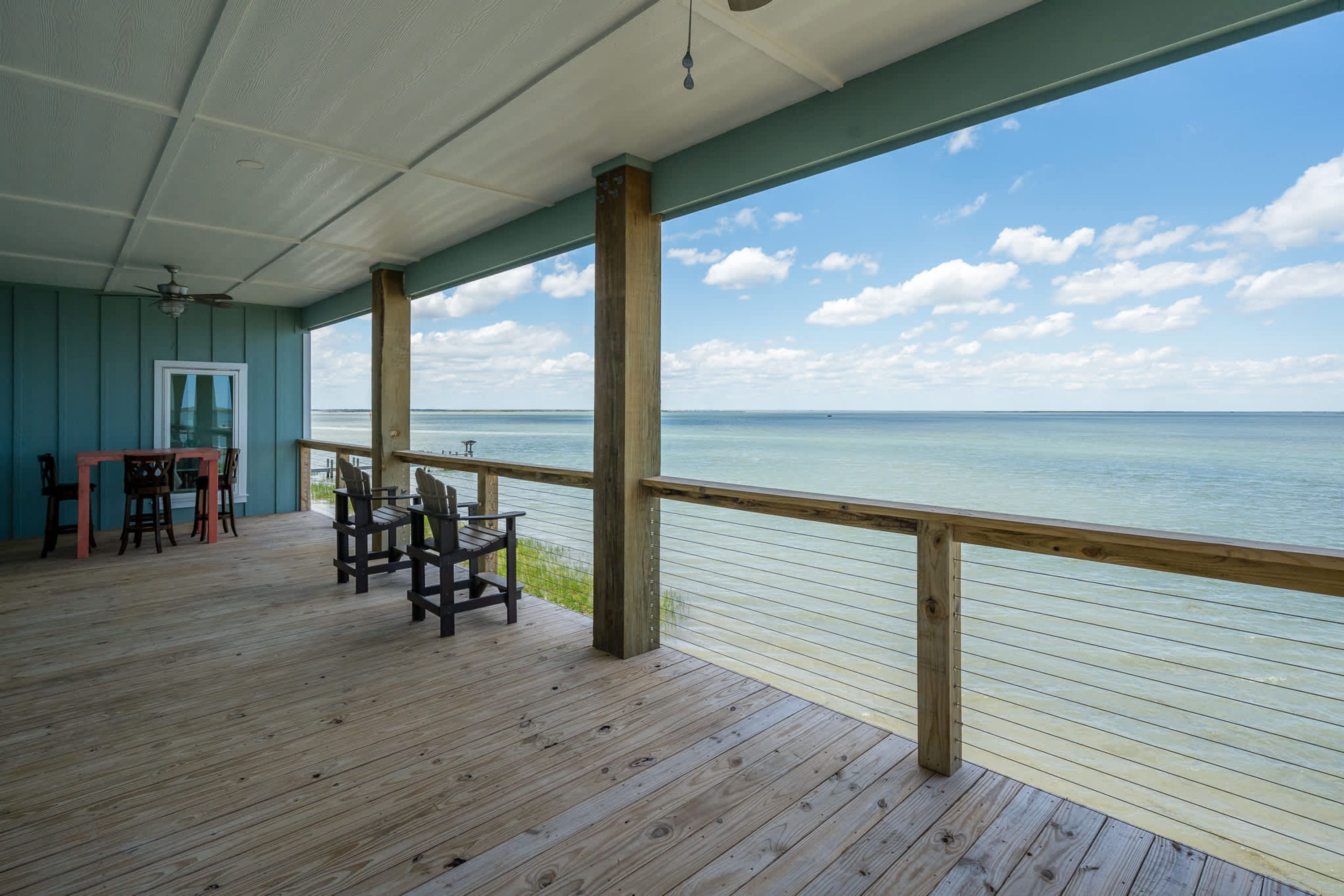 A covered deck with a view of the sea, wooden chairs, a table, and a door.