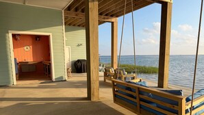 A wooden swing chair with cushions, a wooden deck, a covered grill, and a view of the water. - Waterfront Getaway With Pier Access (Aransas Pass)