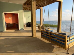A wooden swing chair with cushions, a wooden deck, a covered grill, and a view of the water.