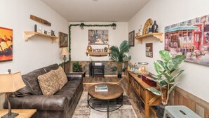 Ein gemütliches Wohnzimmer mit Ledersofa, Couchtisch und Kamin. An Wandschränken befinden sich dekorative Gegenstände und über dem Kamin hängt ein großes Gemälde.