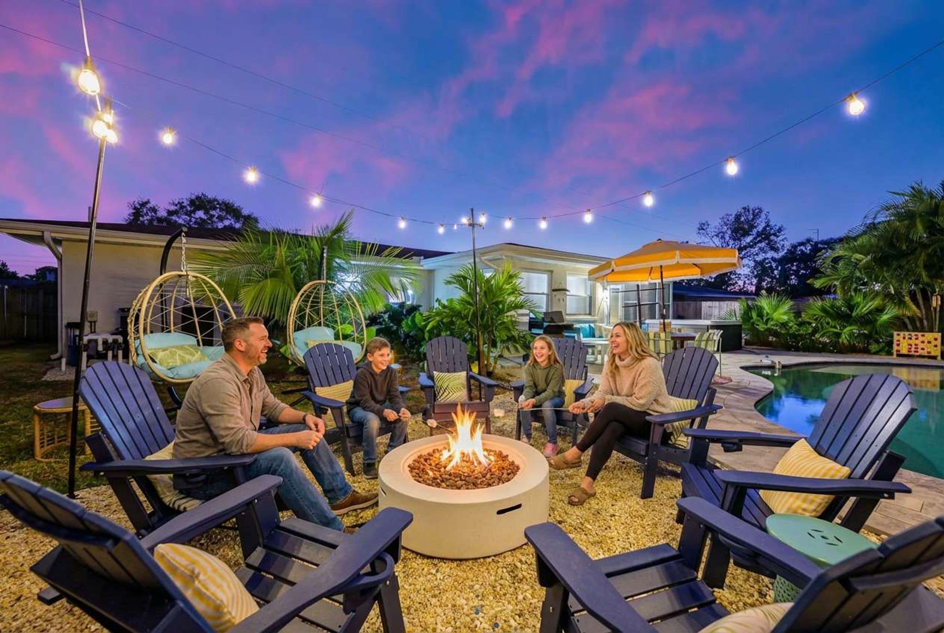 Un grupo de personas sentadas alrededor de un brasero en un entorno al aire libre, con una piscina y una casa al fondo.