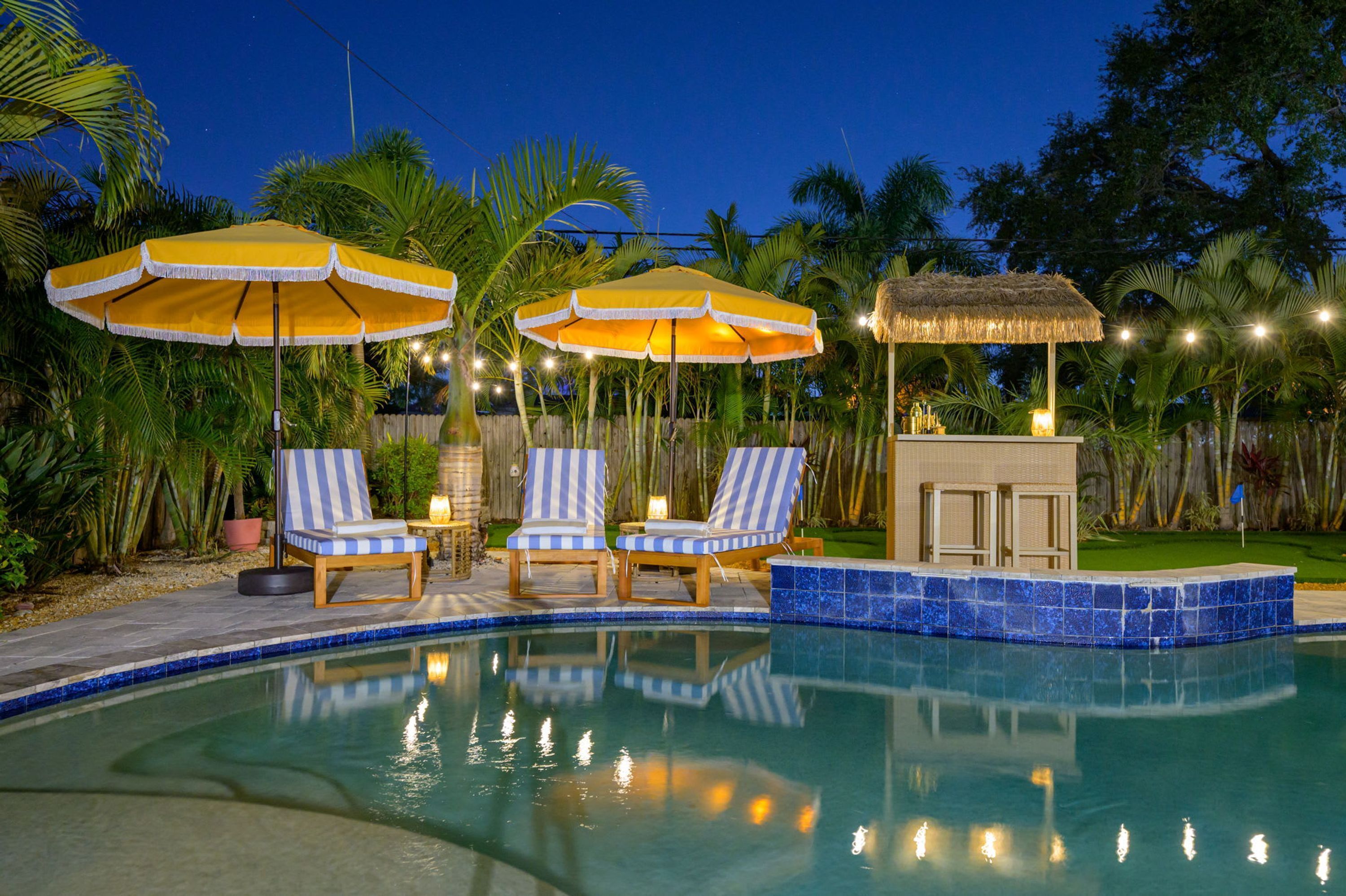 A pool area with lounge chairs and umbrellas, surrounded by palm trees and a thatched structure.