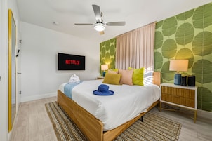 Quarto com cama, ventilador de teto, TV na parede exibindo 'NETFLIX' e mesa de cabeceira com luminária e alto-falante.