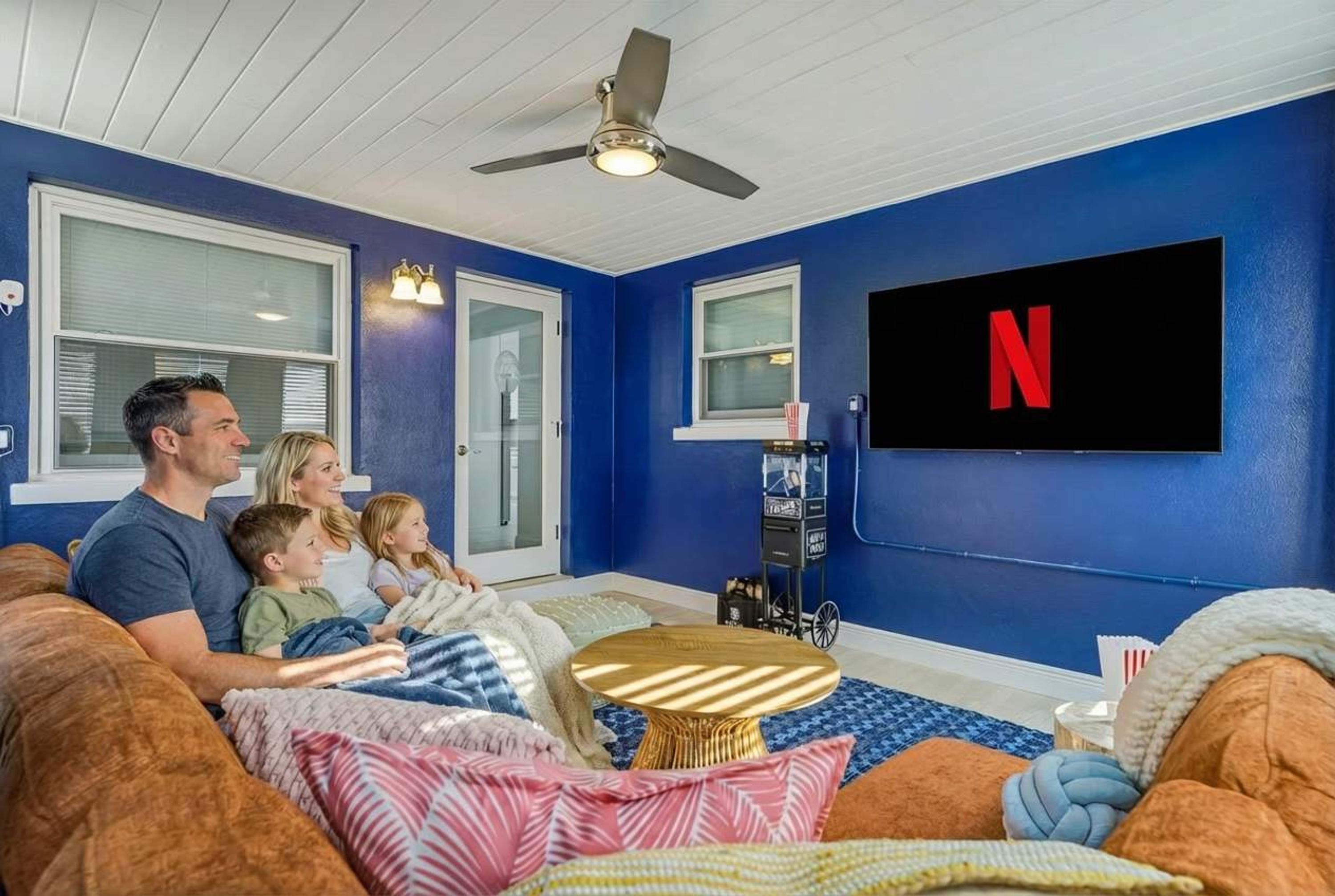 A family watching TV in a room with blue walls, a ceiling fan, and a flat-screen TV displaying the Netflix logo.
