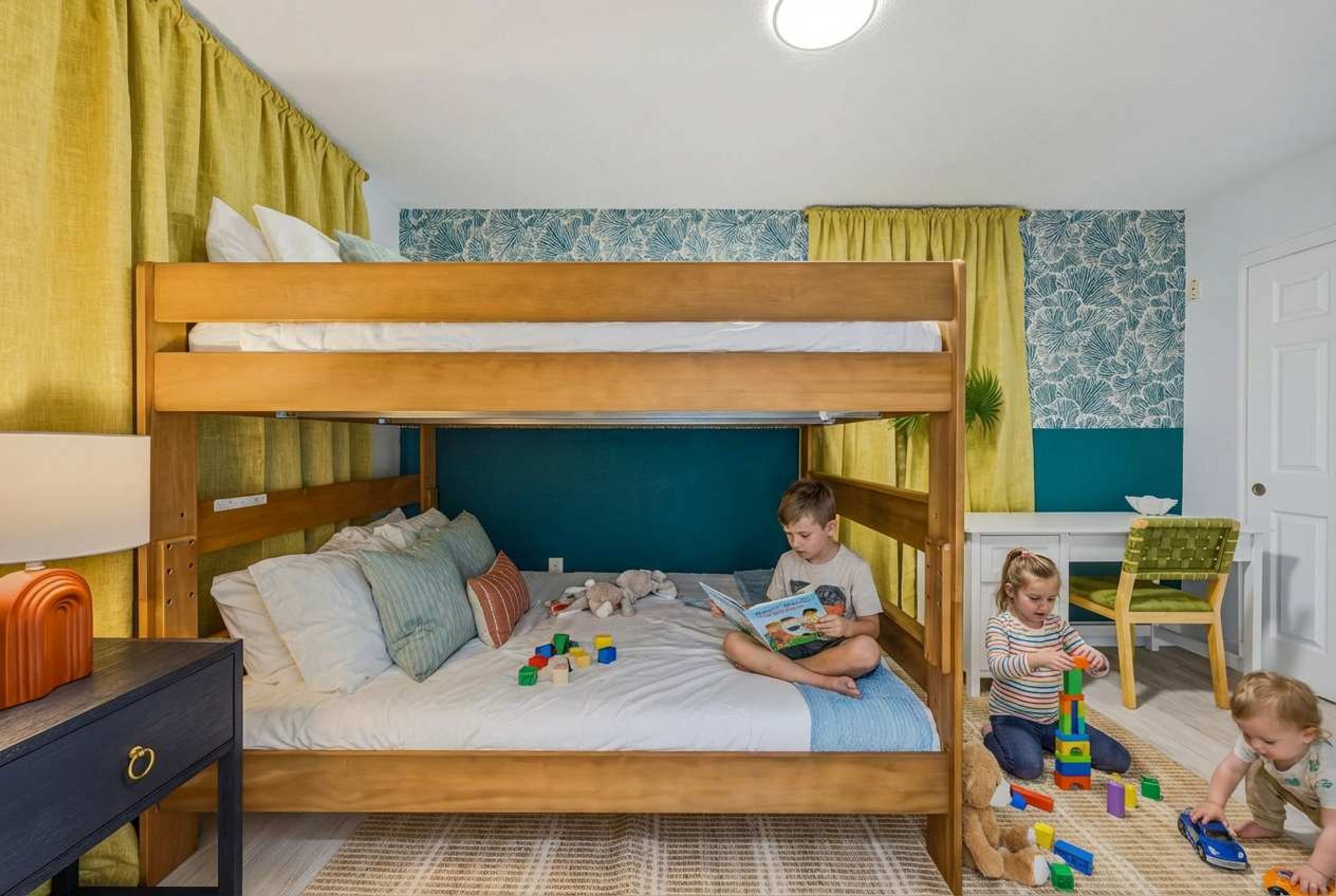 A bunk bed with pillows and toys, a desk with a chair, and a window with curtains.
