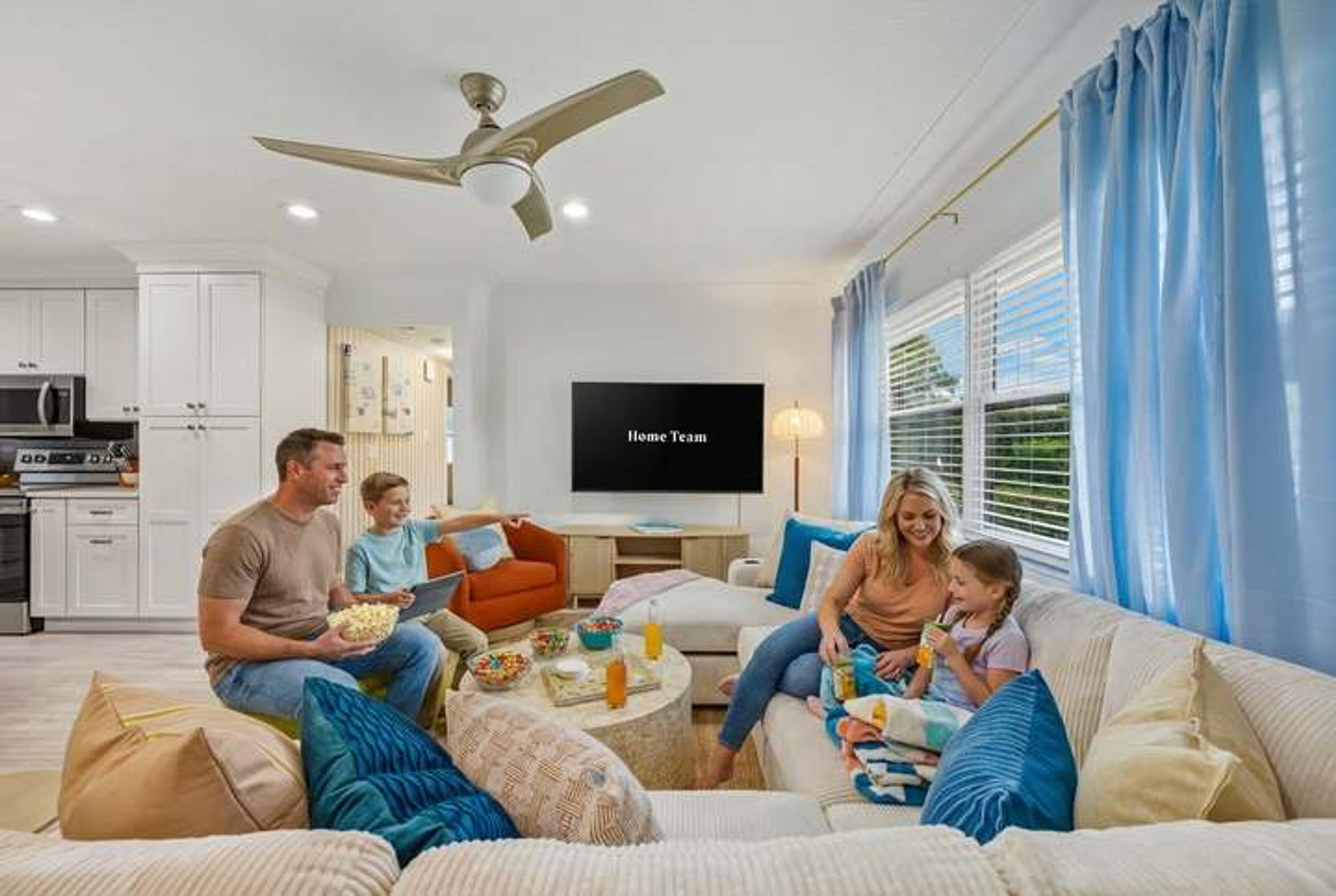 Un salon moderne avec un téléviseur à écran plat, un ventilateur de plafond et une cuisine en arrière-plan.