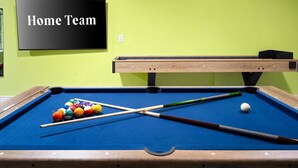 Een pooltafel met ballen en stokken, een 'Home Team' bord en een houten tafel tegen een groene muur.